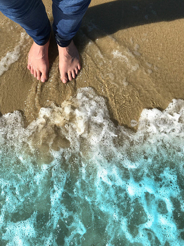 白色的英国男人的脚和紧身的蓝色牛仔牛仔裤在金色的沙滩上被海浪拍打着的海岸线弄湿，用脚趾赤脚划水，夏天的海滨脚印站在海滩上的泡沫/泡沫潮